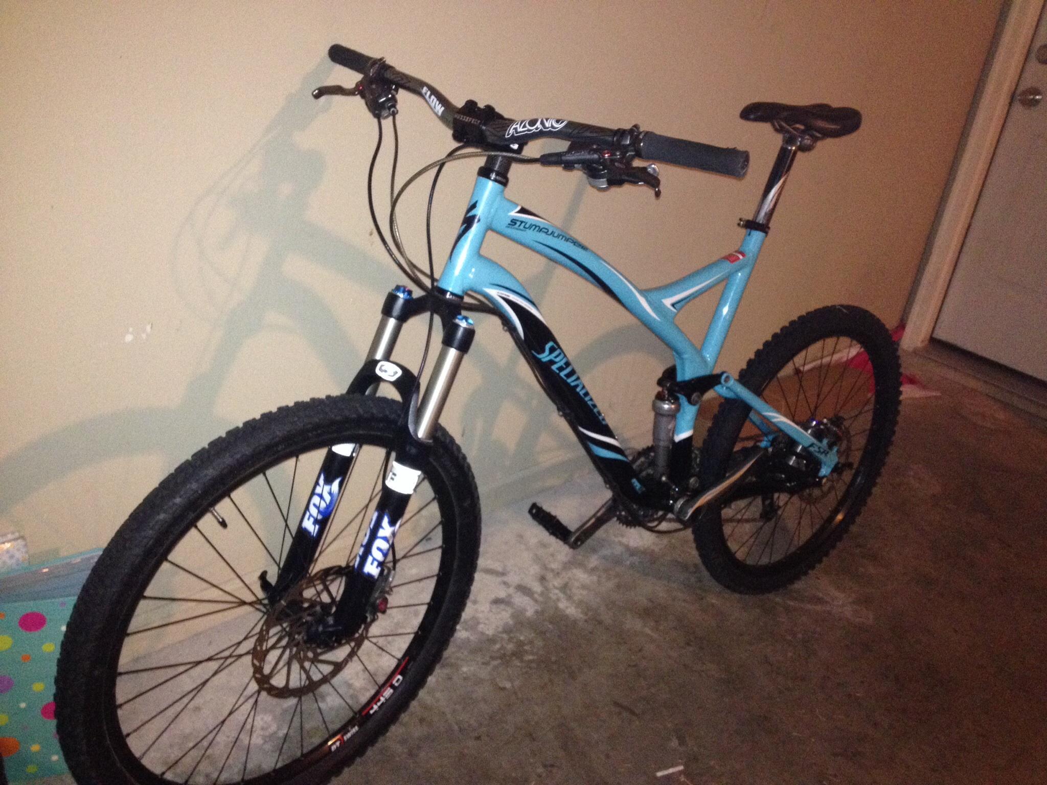 Specialized Stumpjumper FSR Expert: A blue mountain bike is leaning against a wall, showcasing its front suspension forks and knobby tires. The bike features a black and blue design, with visible branding on the frame and tires. The floor has a concrete finish, and there is a door visible in the background.