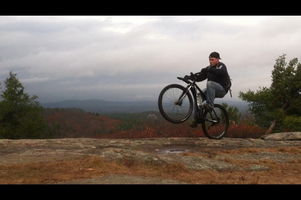 A cyclist performing a wheelie on a mountain bike on a rocky surface, with a scenic view of rolling hills and cloudy skies in the background. The surroundings feature trees and autumn foliage, suggesting a natural outdoor setting. Franklin Falls mountain bike trail.