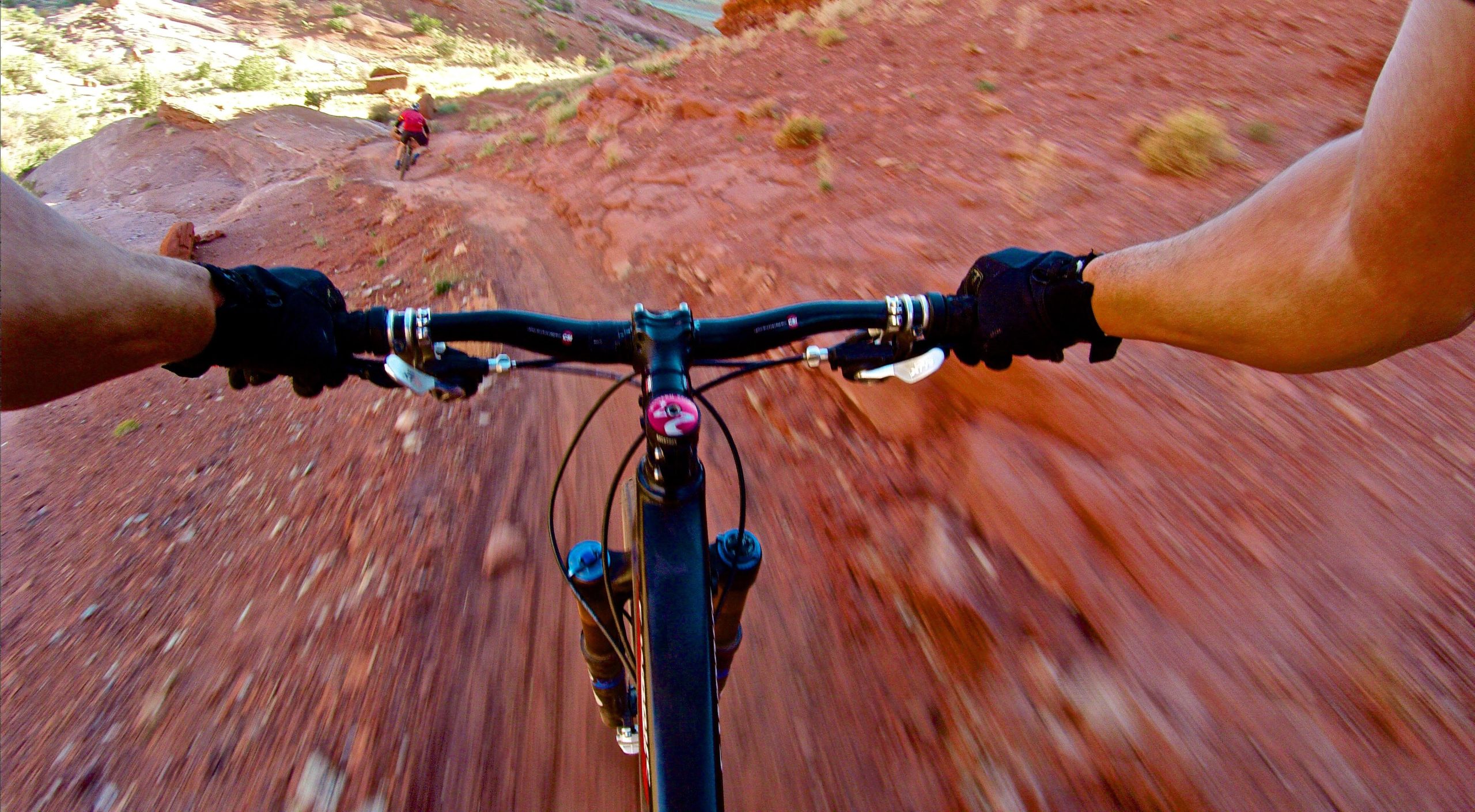 A cyclist's perspective captures the handlebars of a mountain bike while navigating a winding, reddish dirt trail. The background shows another rider in a red shirt on a similar path, surrounded by rugged terrain and greenery. The image conveys a sense of speed and adventure in a natural outdoor setting. Moab Brand Trails mountain bike trail.