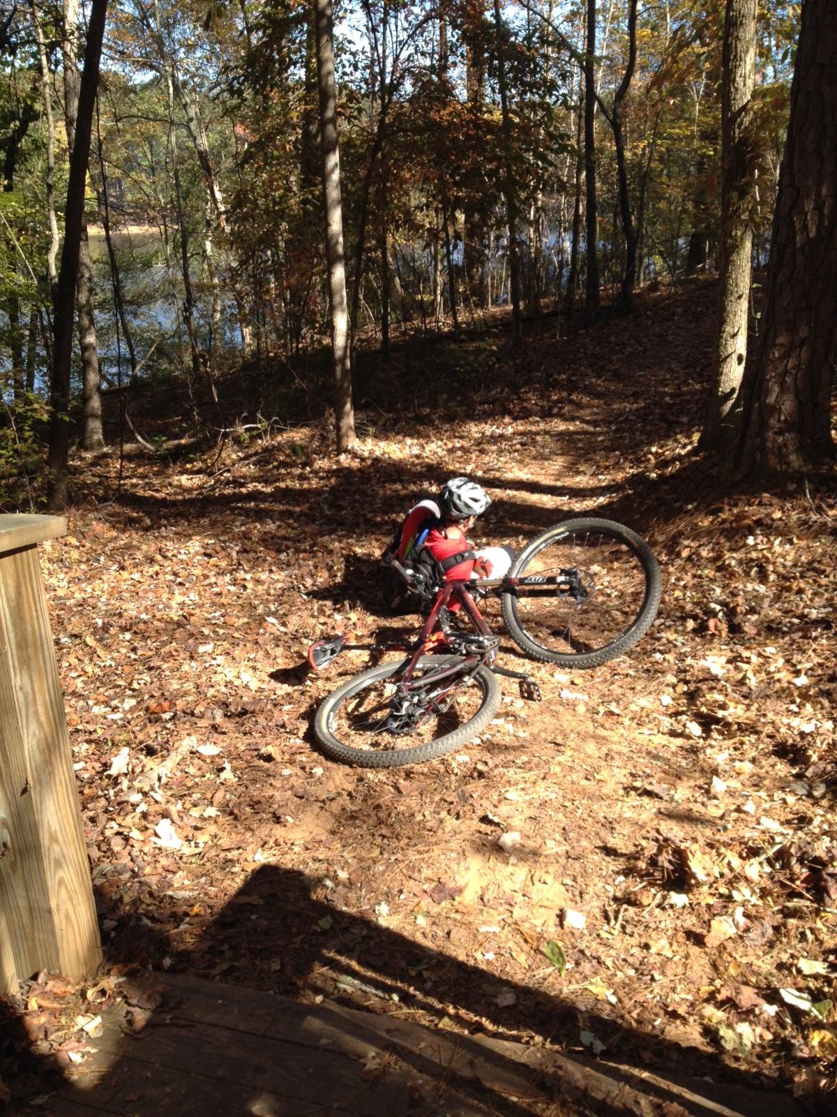A fallen mountain bike lies on a dirt path covered with autumn leaves, near a wooded area by a lake. In the background, trees with colorful foliage can be seen, and the sunlight filters through the branches, creating a serene outdoor atmosphere. Dauset Trails Nature Center mountain bike trail.