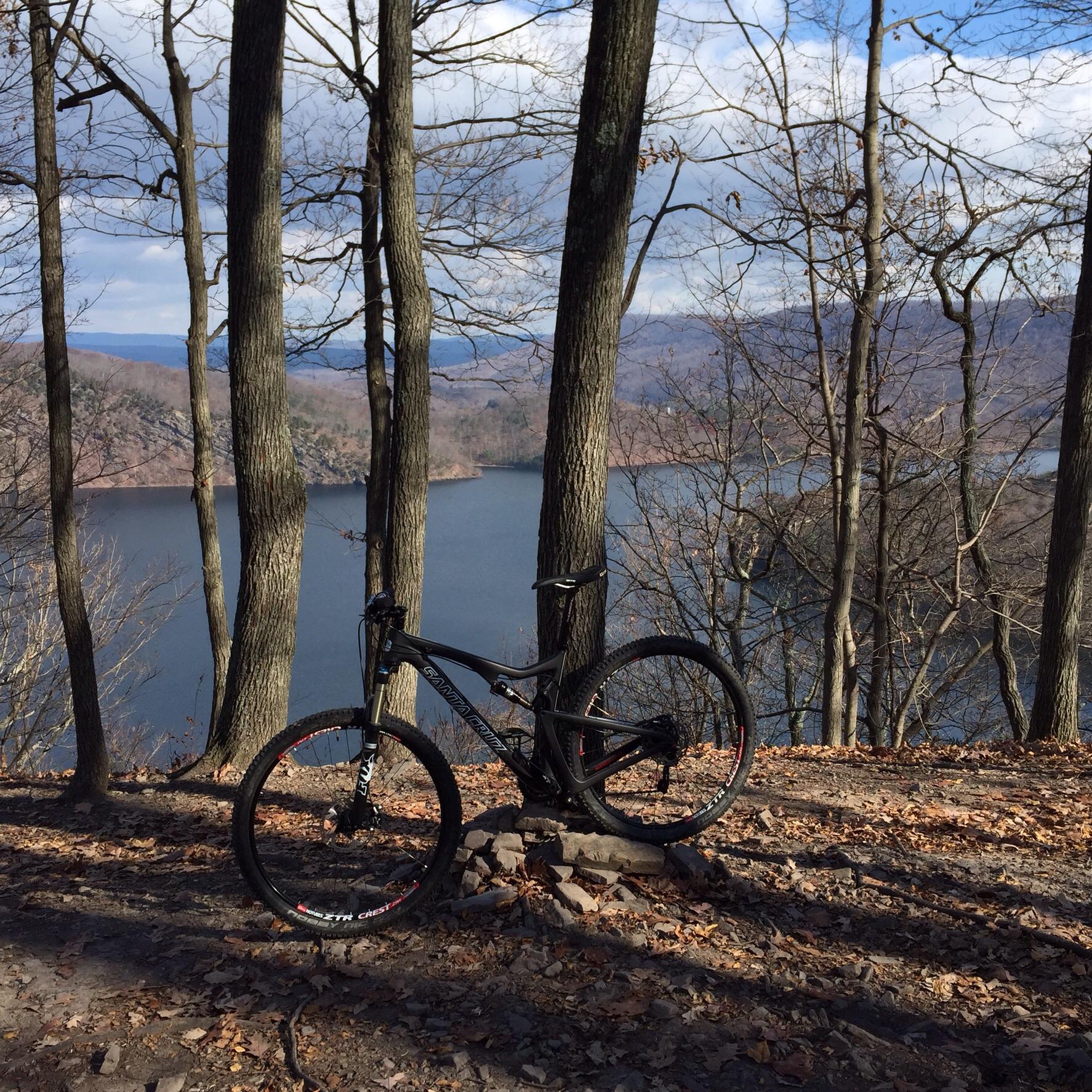 A mountain bike resting against a rock on a wooded trail, with a scenic view of a lake and distant hills in the background. The trees are bare, suggesting an early spring or late fall setting. Allegrippis Trails mountain bike trail.