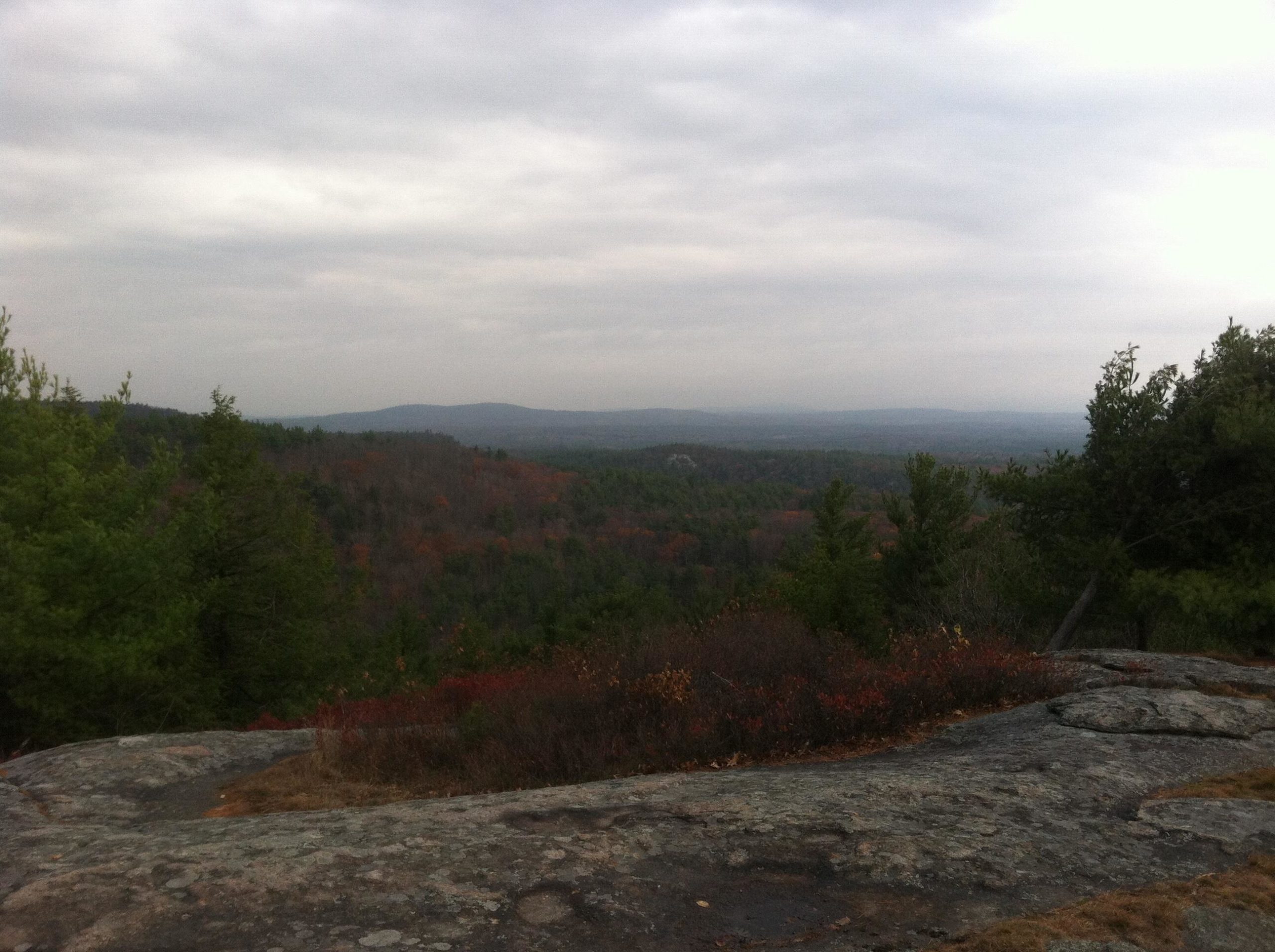 A panoramic view of a hilly landscape under a cloudy sky, showcasing a mix of evergreen trees and deciduous trees with autumn foliage in shades of green and red. The rocky foreground adds texture to the scene, while distant mountains create a sense of depth. Franklin Falls mountain bike trail.