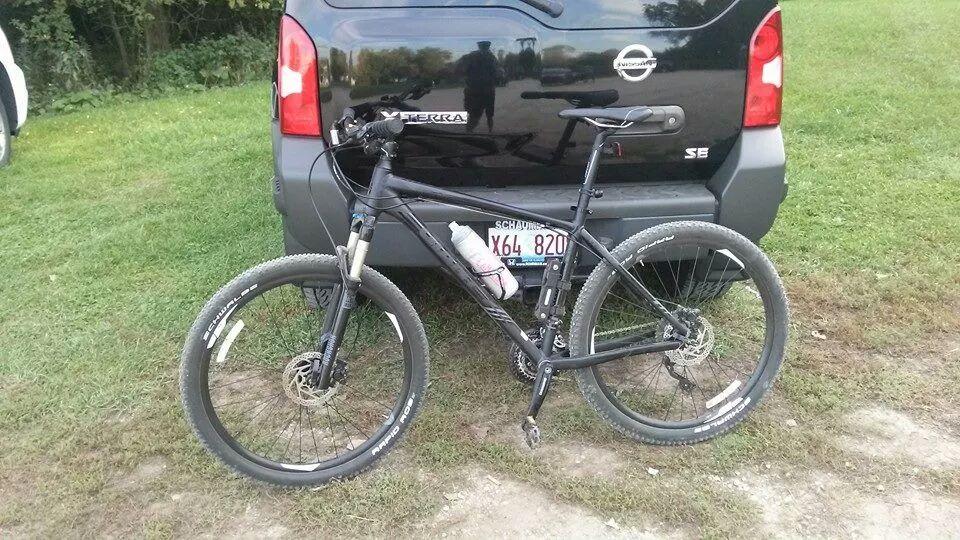 Giant Talon 27.5: A black mountain bike with knobby tires is parked on grass next to the rear of a Nissan SUV. A water bottle is attached to the bike frame. The background includes a tree line and part of a gravel path.