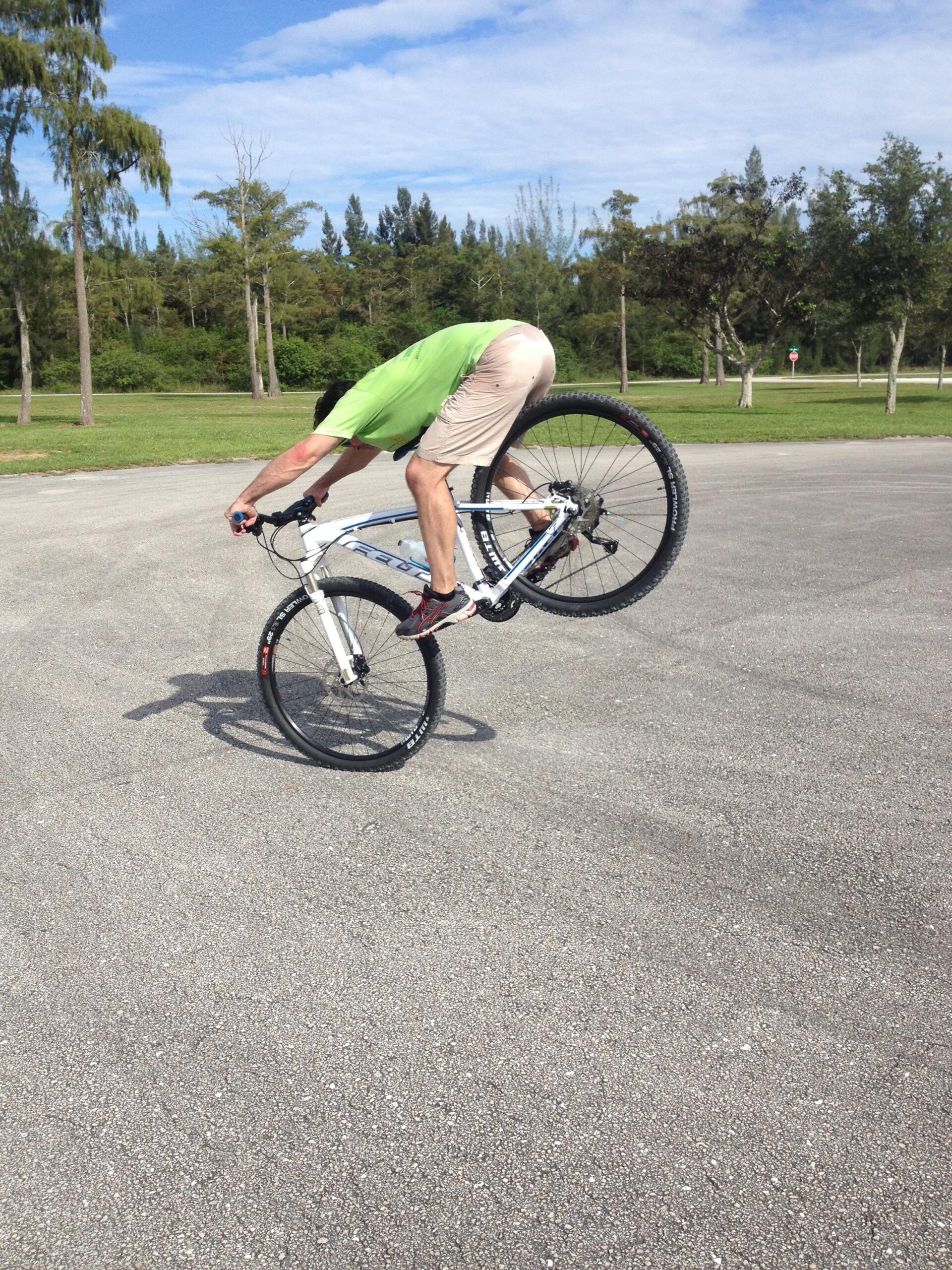 Felt Nine 50: A person riding a mountain bike is performing a wheely, leaning forward over the front wheel while balancing on the rear tire. The setting is an outdoor area with grassy terrain and scattered trees under a partly cloudy sky.