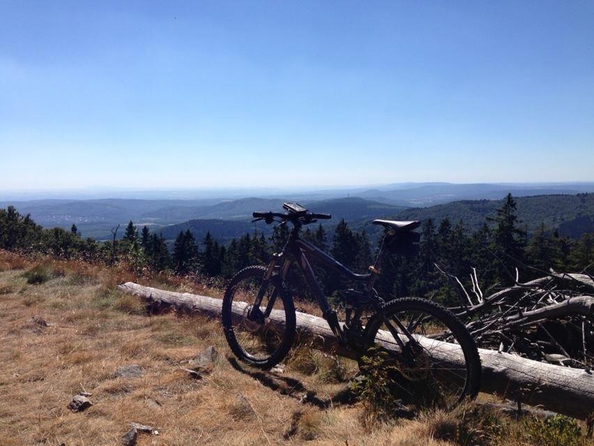 Mountain bike resting on a log in a grassy clearing, with a panoramic view of rolling hills and distant mountains under a clear blue sky. Taunus mountain bike trail.