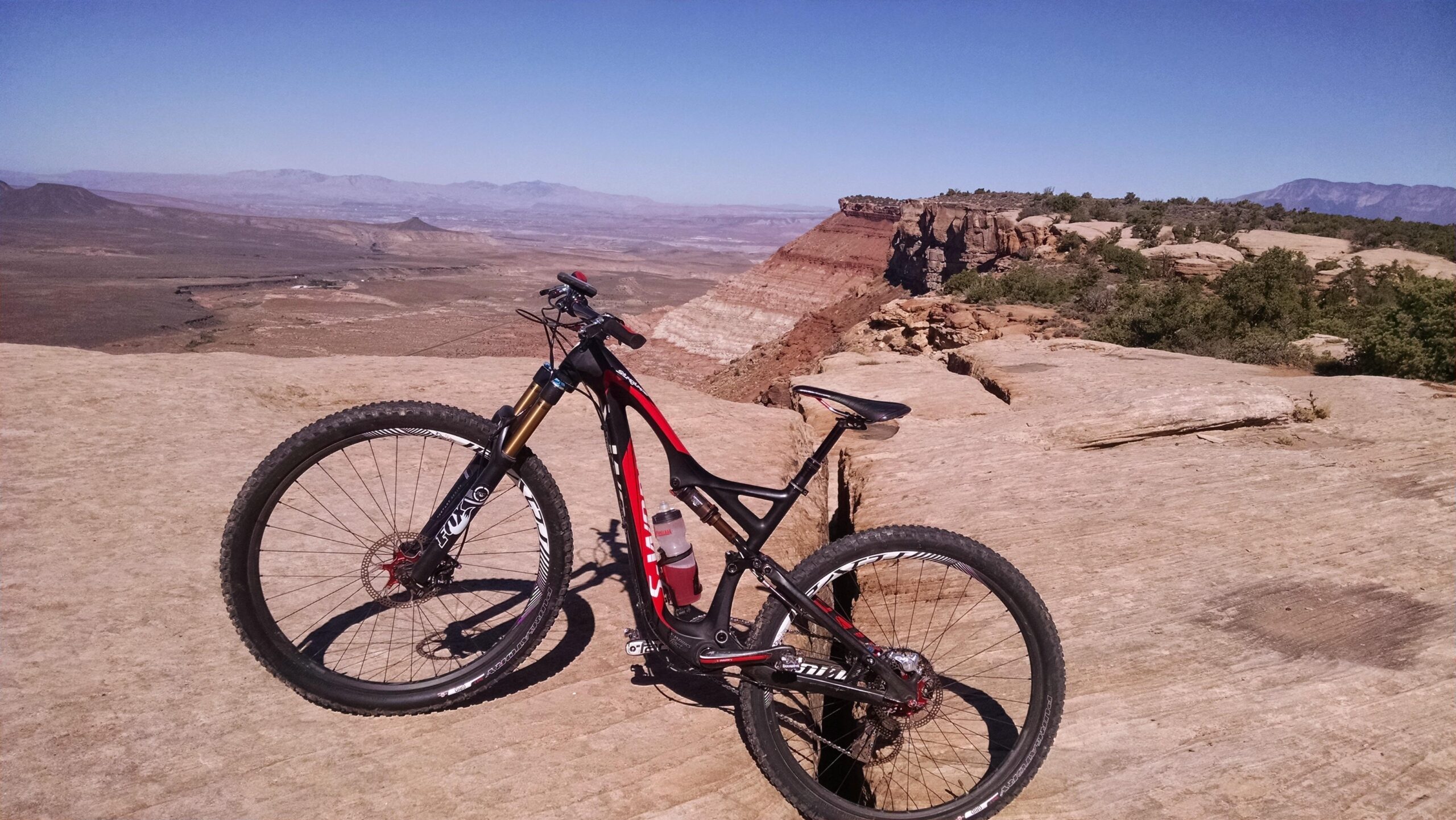 Specialized S-Works Stumpjumper FSR 29: A mountain bike is parked on a rocky plateau overlooking a vast, arid landscape with distant mountains and a clear blue sky. The bike features a black and red frame, and a water bottle is attached to its side. The scenery includes layered rock formations in the background, adding to the rugged outdoor setting.