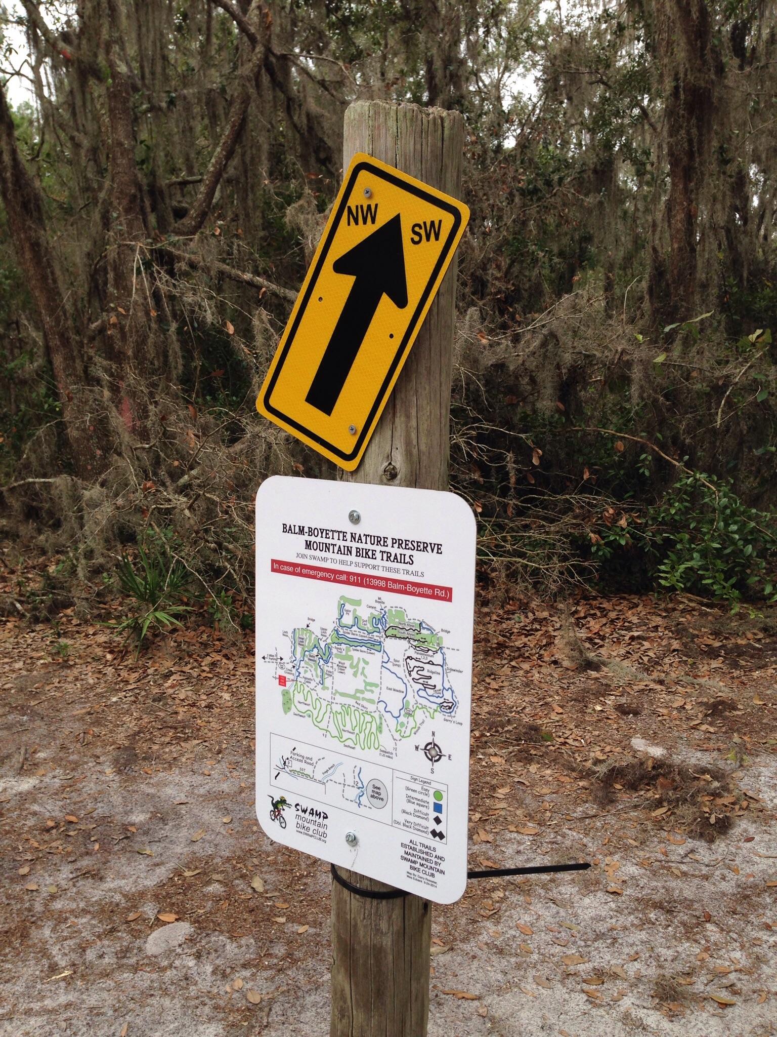 A wooden post supporting a directional sign pointing straight ahead, labeled "NW" and "SW," alongside a map sign detailing the Balm Boyette Nature Preserve mountain bike trails, with trees and foliage in the background. Balm Boyette Scrub Preserve mountain bike trail.