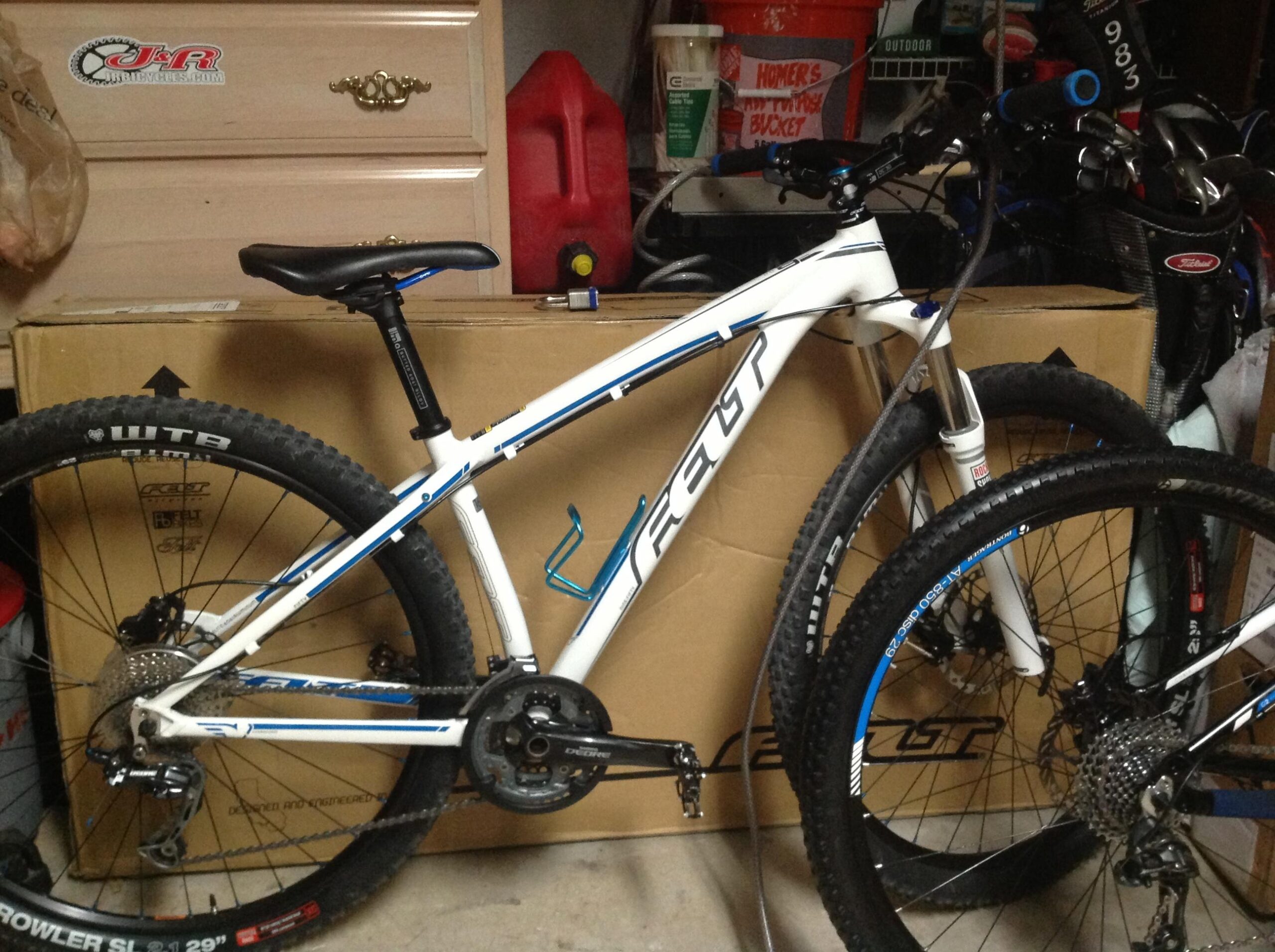 Felt Nine 50: A white mountain bike with blue accents is positioned next to a cardboard box in a garage setting. The bike features thick tires, a visible gear system, and a water bottle holder. Behind the bike, various items including a gas can, buckets, and golf clubs are partially visible.