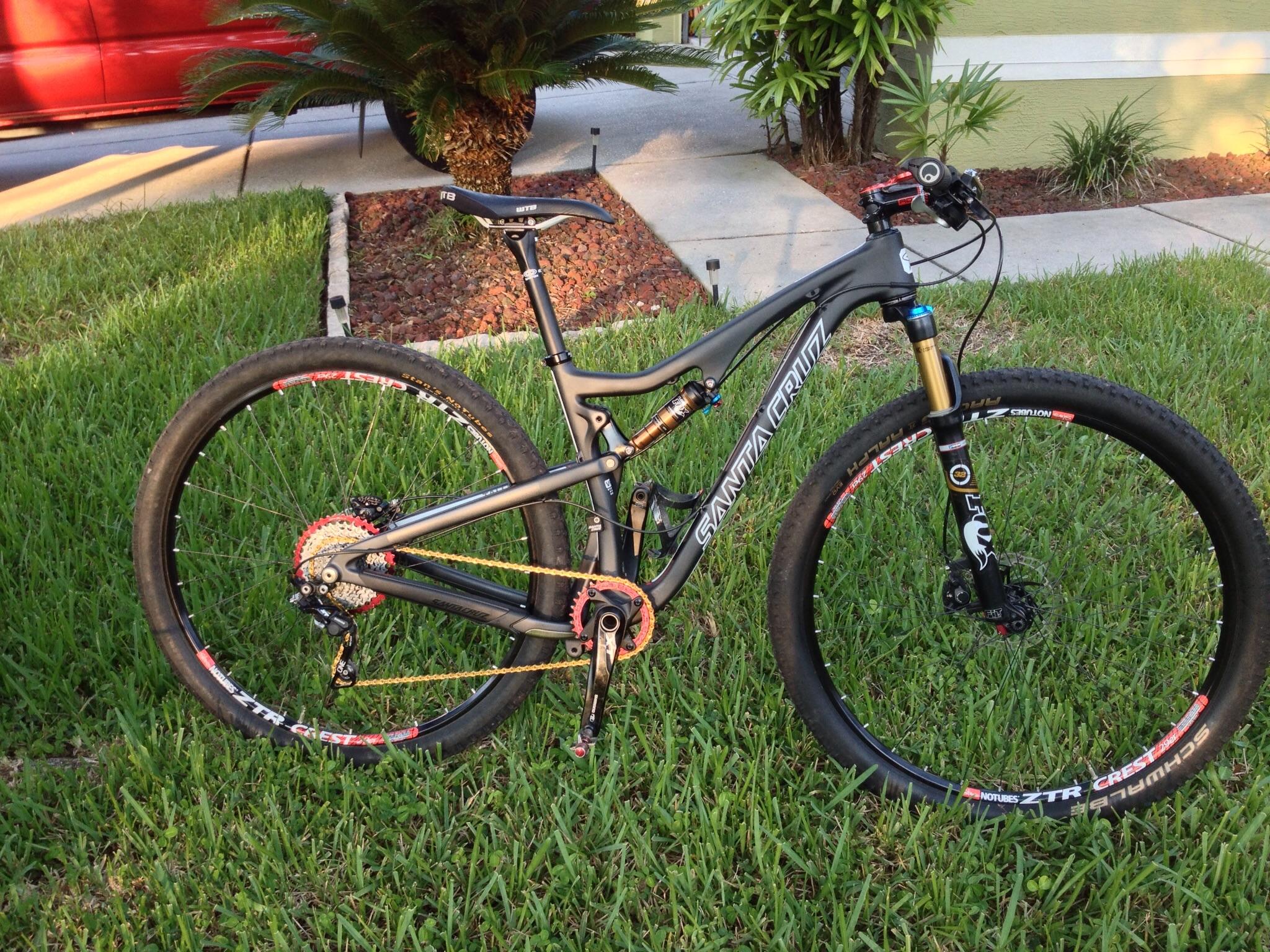 Santa Cruz Tallboy Carbon: A black Santa Cruz mountain bike parked on green grass, featuring large tires, a gold fork, and various colored components. The bike is positioned at an angle, showcasing its frame design and gear setup. In the background, a well-maintained lawn and landscape add depth to the scene.