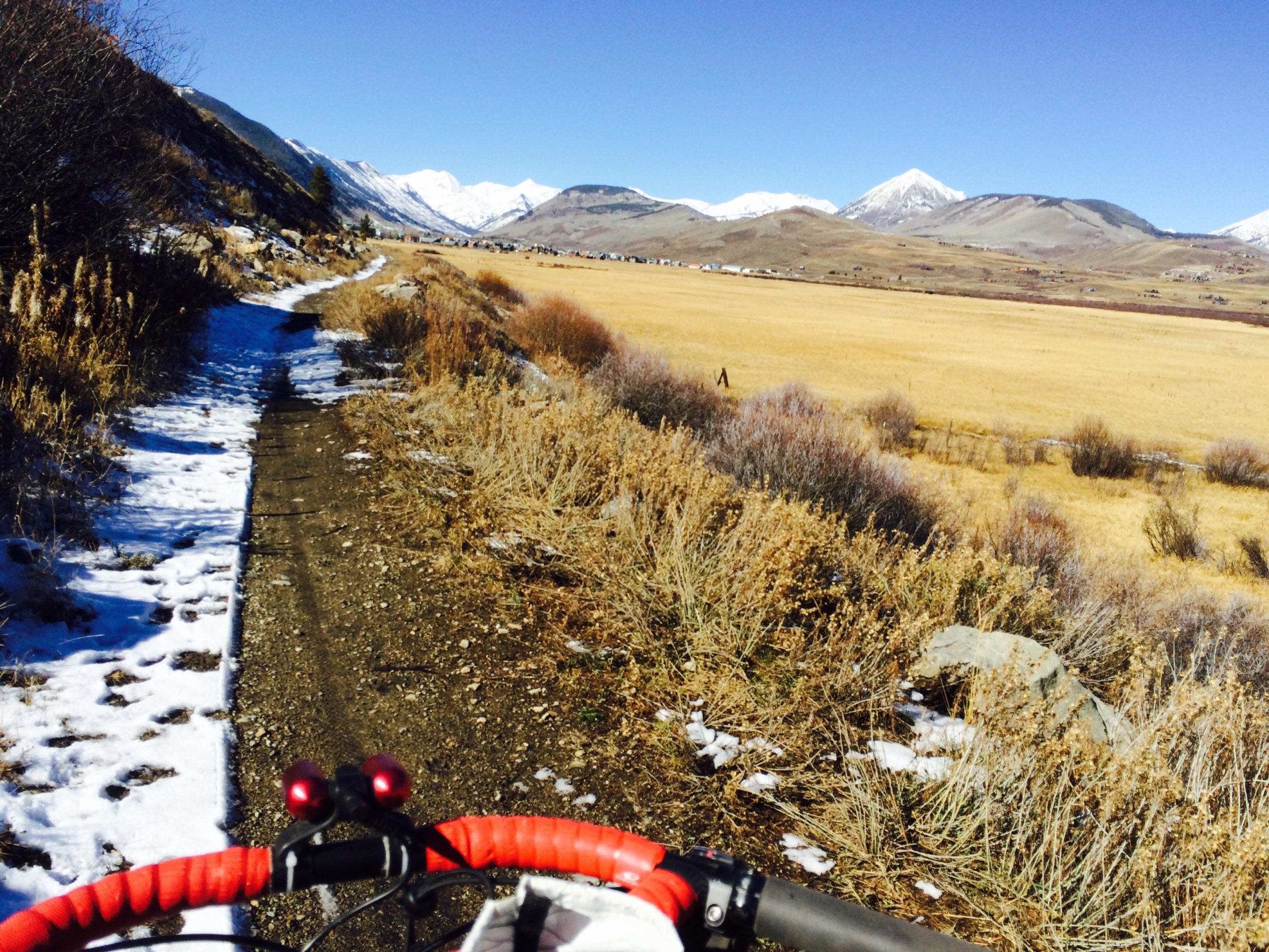 A view of a biking path winding through a mountainous landscape, with snow on one side and dry grasses on the other. Snow-capped mountains rise in the background against a clear blue sky, while a golden field stretches out in the foreground. The handlebars of a bike, adorned with red grips, are visible in the bottom corner of the image. New Deli Trail mountain bike trail.