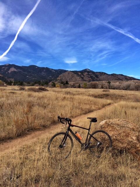 Giant Revolt 1: A black bicycle leaning against a large rock on a dirt path surrounded by tall grass, with mountains and a clear blue sky in the background, featuring streaks of clouds.