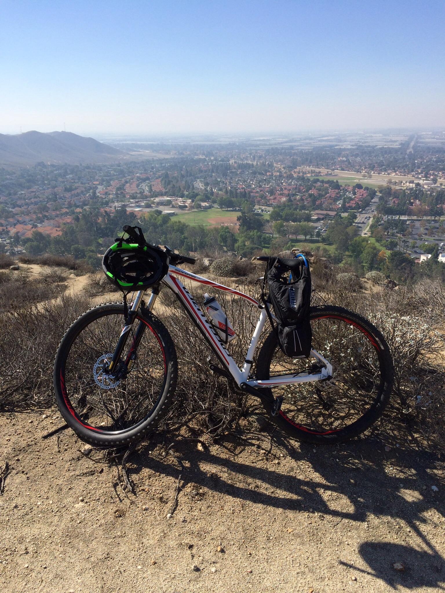 Specialized Rockhopper Comp 29: A mountain bike parked on a rocky path overlooking a scenic valley. The landscape includes homes, trees, and fields, with mountains in the background under a clear blue sky. A green and black helmet is attached to the bike, along with a black backpack.