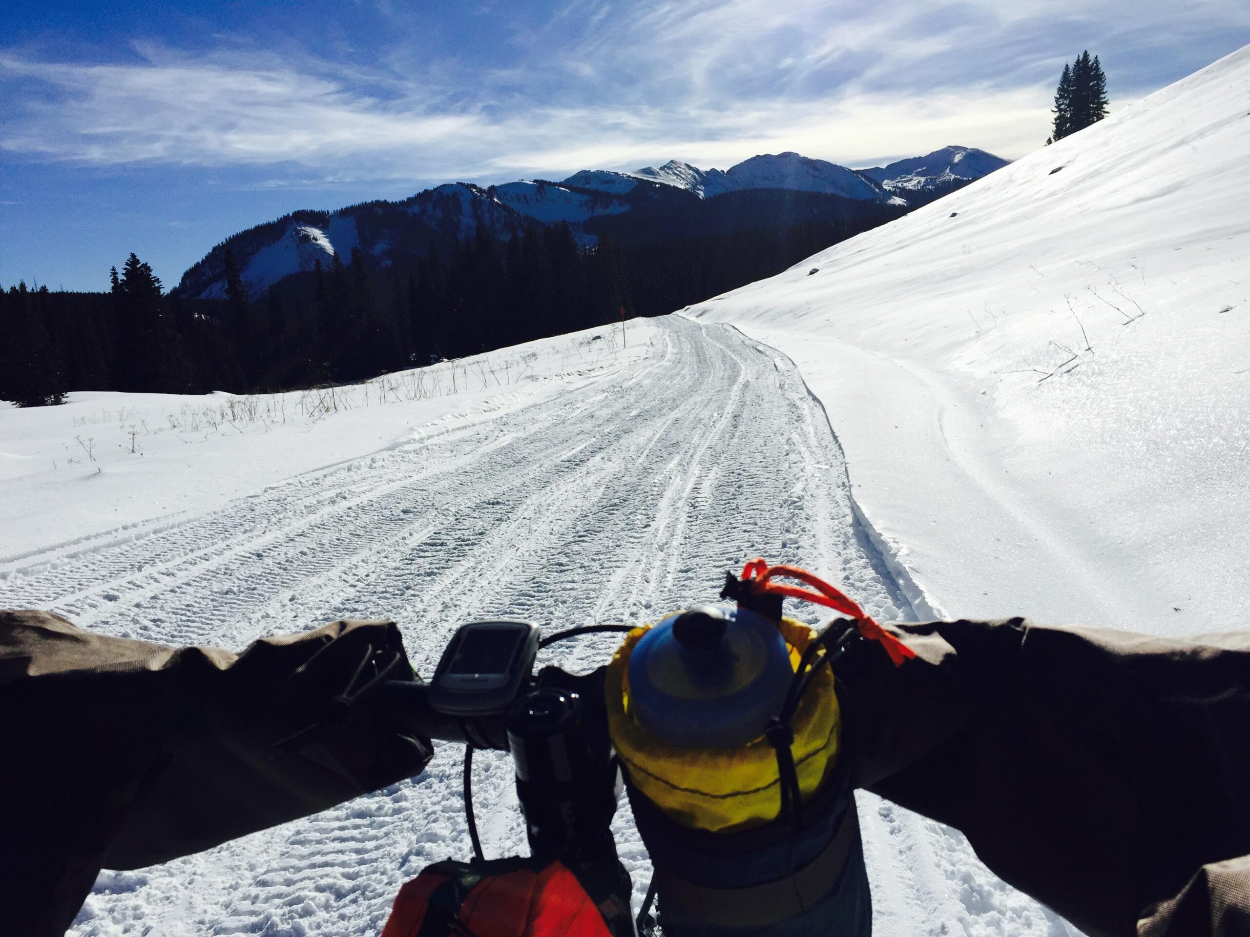A view from the handlebars of a bike on a snowy mountain trail, with a clear blue sky and rugged peaks in the background. The bike is equipped with a yellow water bottle, and snow-covered grounds feature tire tracks indicating prior use. Kebler Pass Road mountain bike trail.