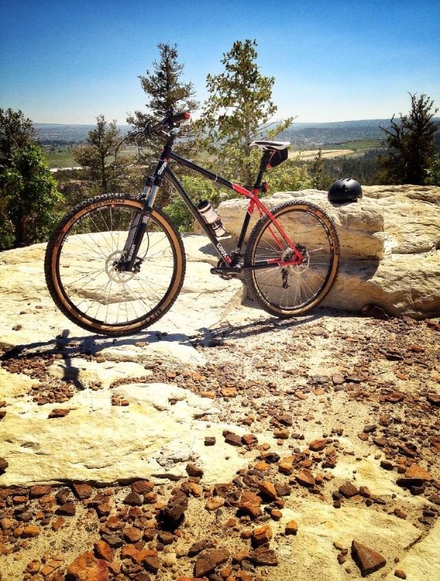 Charge Cooker 1: A mountain bike with a black and red frame is parked on a rocky outcrop, with a helmet resting nearby. The background features a clear blue sky and a distant view of hills and valleys, indicating a scenic outdoor location.