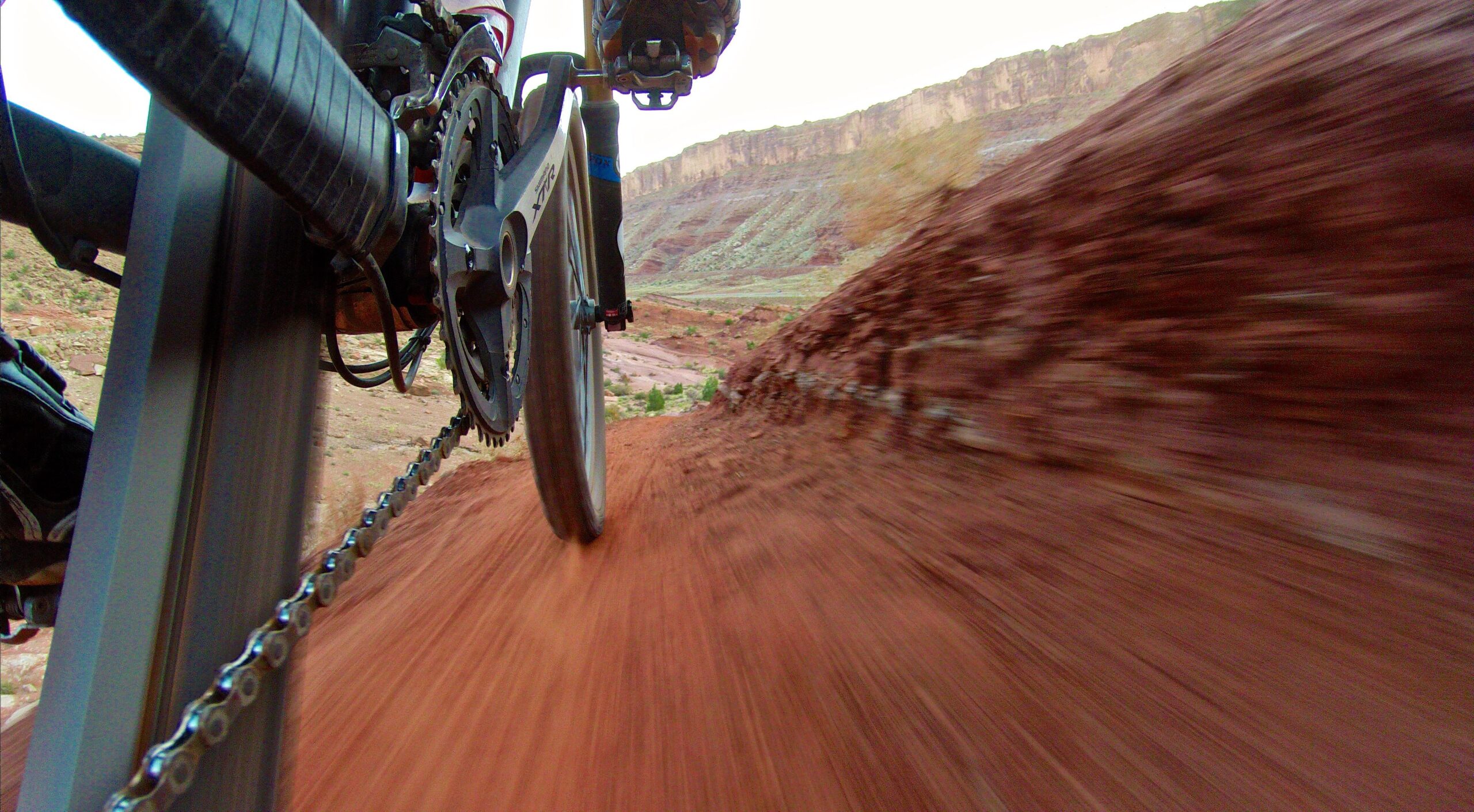 Close-up view of a mountain bike wheel and chain as it rides along a reddish dirt trail, with scenic cliffs in the background, suggesting speed and motion. Moab Brand Trails mountain bike trail.