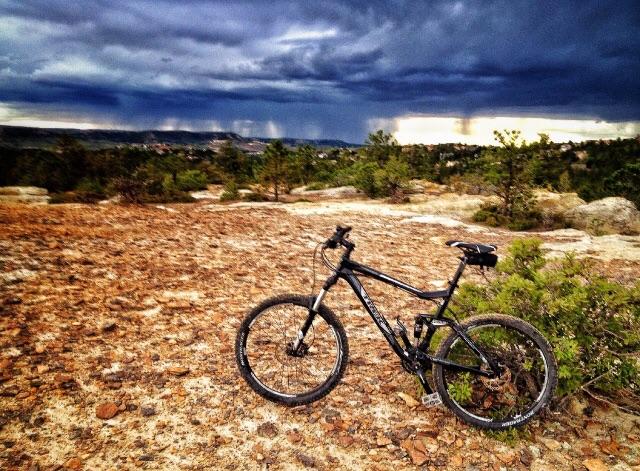 Trek Fuel EX 6 26: A mountain bike resting on rocky terrain with a backdrop of dark, stormy clouds and distant rain falling on the horizon. Green shrubs and trees are scattered around the landscape, creating a rugged outdoor scene.