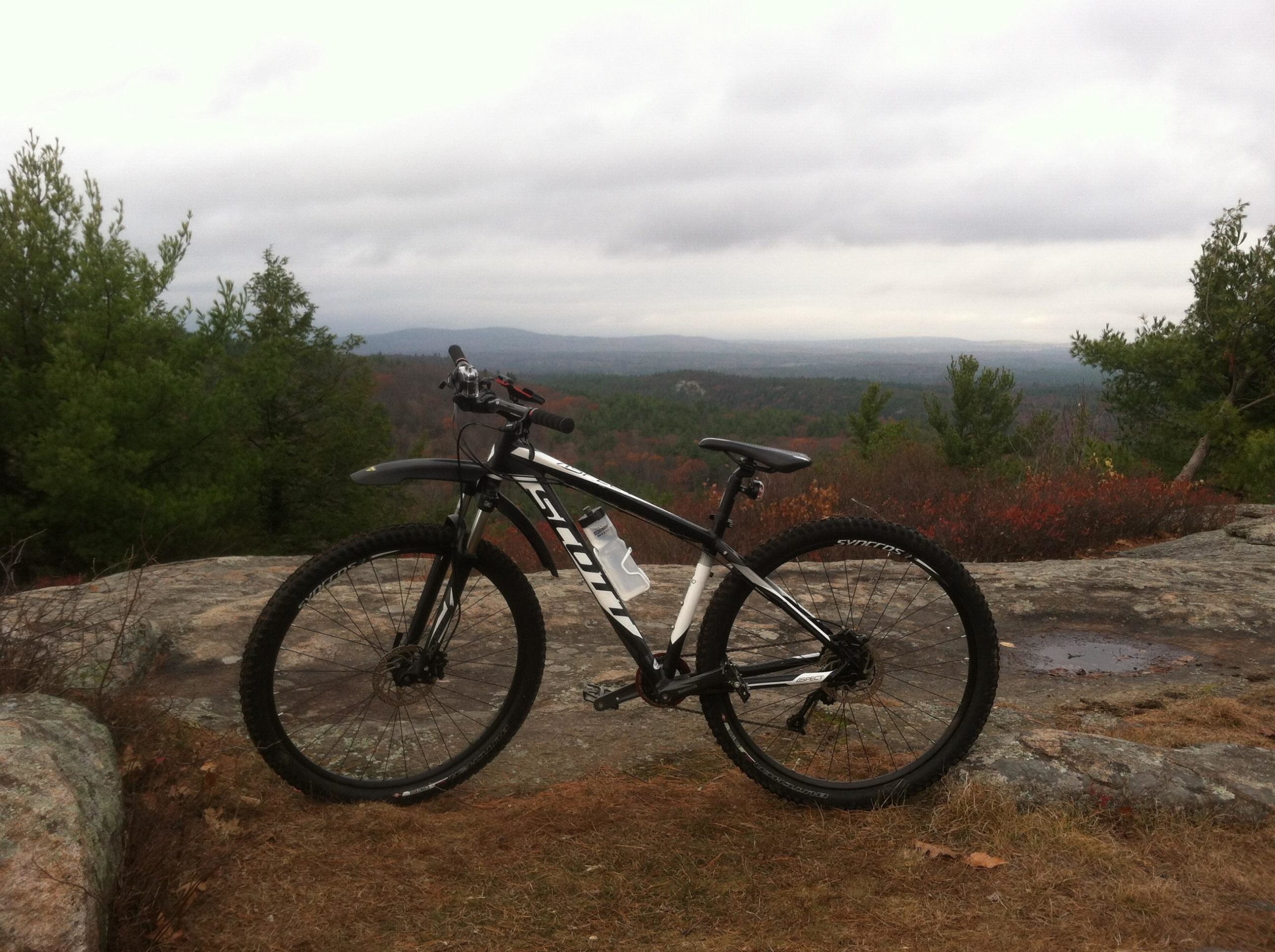 Mountain bike parked on a rocky ledge with a scenic view of rolling hills and forests under a cloudy sky. Franklin Falls mountain bike trail.
