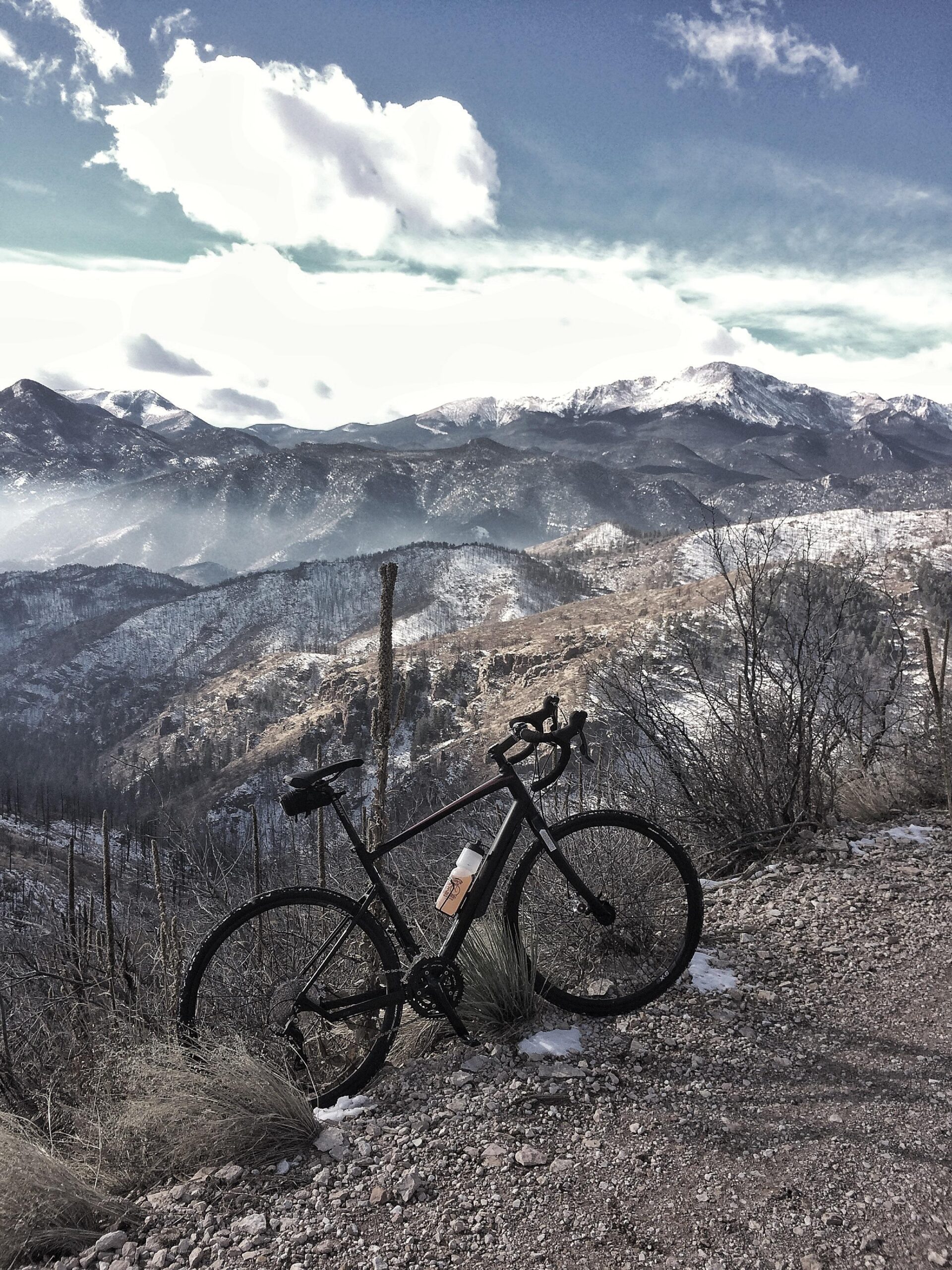 Giant Revolt 1: A black bicycle with a water bottle is parked on a gravel path, overlooking a mountainous landscape. Snow-capped peaks are visible in the background, with a mix of rocky hills and sparse vegetation in the foreground. The sky is partly cloudy, creating a serene and picturesque outdoor scene.