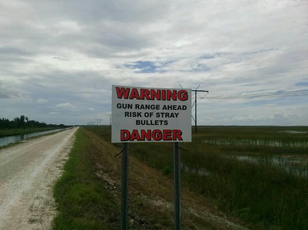 A warning sign indicating the presence of a gun range ahead, with alerts about the risk of stray bullets. The sign is prominently displayed alongside a dirt road, surrounded by grassy fields and a cloudy sky. Conservation Levee Greenway mountain bike trail.