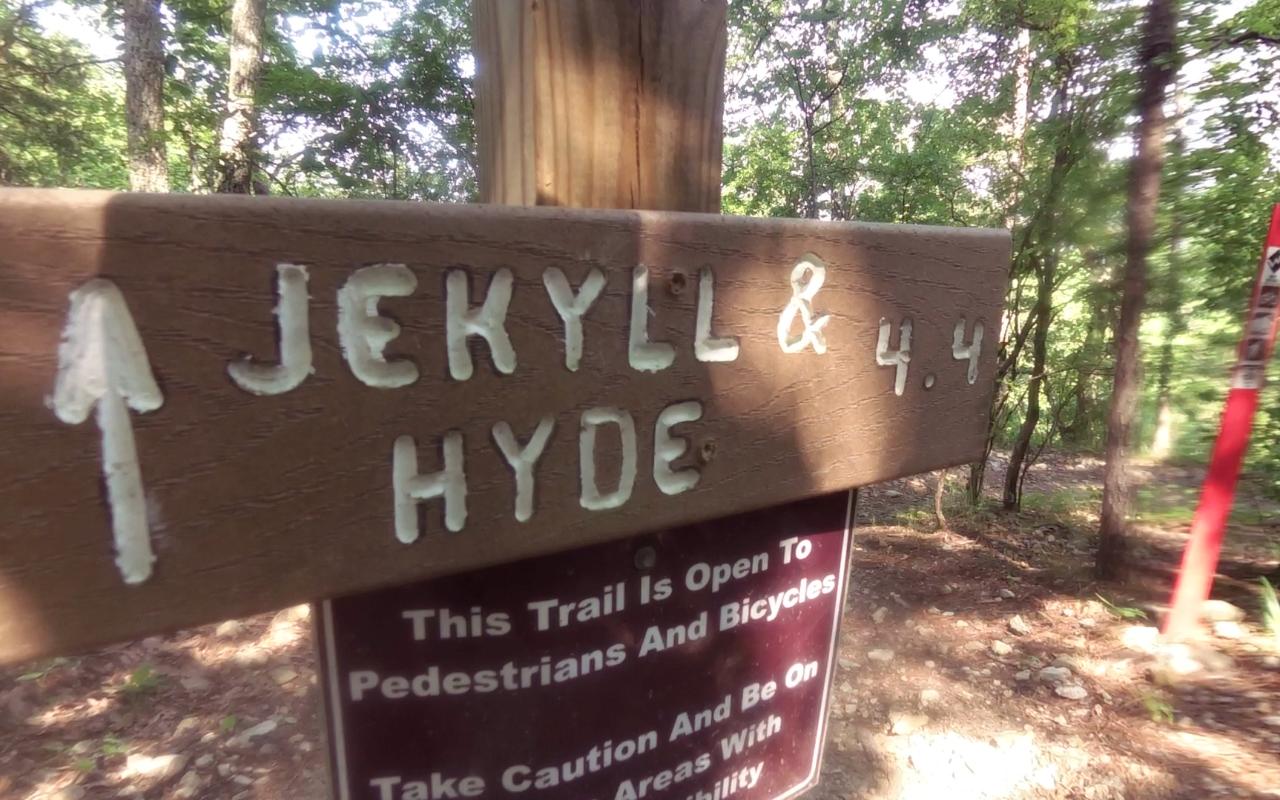 A wooden trail sign indicating "Jekyll & Hyde" with an arrow pointing upwards, alongside a distance of 4.4 miles. In the lower section, a safety notice reads: "This trail is open to pedestrians and bicycles. Take caution and be on areas with instability." The background features trees and a natural setting. Oak Mountain State Park Bump Trail mountain bike trail.