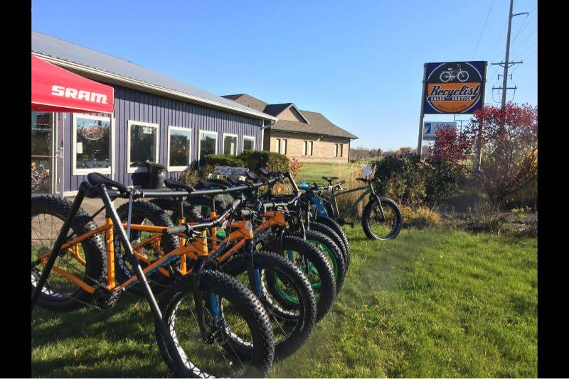 A row of colorful bicycles displayed in front of a bike shop on a sunny day. The shop features a sign with the name "Recyclist" and a visible "Open" sign in the window. Lush green grass and some shrubs are visible in the foreground, while a clear blue sky is overhead.