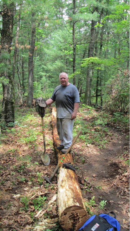 A man standing on a fallen log in a wooded area, holding a shovel and a hat. The surrounding forest is lush with green trees and foliage. A backpack is placed on the ground nearby, and the scene conveys a sense of outdoor activity or gardening. Russell Mill mountain bike trail.
