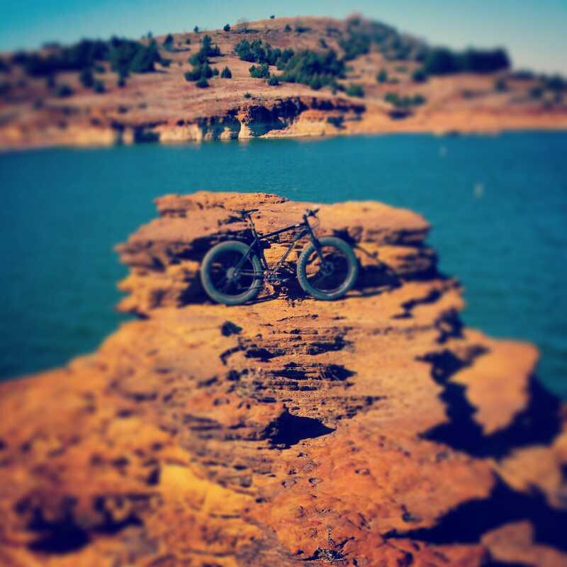 A black fat bike resting on a rocky outcrop by a blue lake, surrounded by a dry, hilly landscape under a clear blue sky. The scene captures a serene outdoor setting ideal for biking adventures. Switchgrass mountain bike trail.