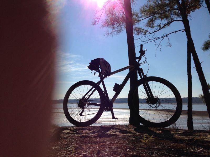 Silhouette of a bicycle leaning against a tree, with a lake and blue sky in the background. The sun is shining, creating a bright glow and reflections on the water. Clear Creek mountain bike trail.