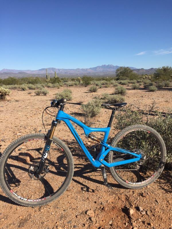A blue mountain bike is parked on a rocky desert landscape, surrounded by sparse vegetation and low shrubs. In the background, faint mountain ranges are visible under a clear blue sky. McDowell Mountain Park mountain bike trail.