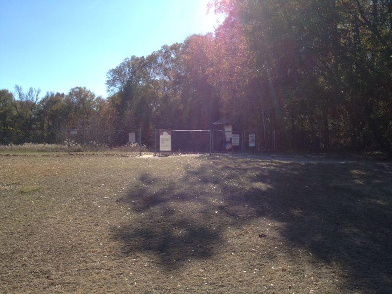 A grassy area with a chain-link fence and a gated entrance to a park or recreational area. Trees are visible in the background, and the scene is brightly lit by sunlight. L.H. Thomson Trails mountain bike trail.