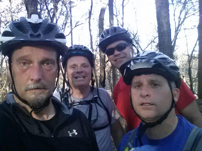 Four men wearing helmets are posing for a selfie in a wooded area. They appear to have just finished a biking adventure, with one person smiling and the others looking more serious. The background features trees and natural greenery, suggesting a vibrant outdoor setting. Harbison State Forest mountain bike trail.