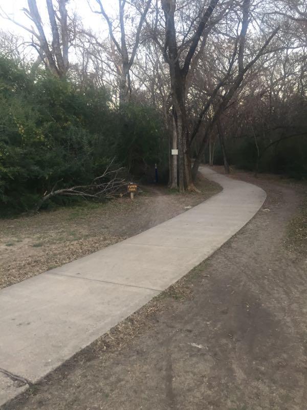 A winding concrete path leading through a tree-lined area, with sparse foliage and bare branches. A wooden sign is visible on the left, partially hidden by vegetation. The scene conveys a quiet, natural setting, with a blend of paved and dirt surfaces. Gateway Park mountain bike trail.
