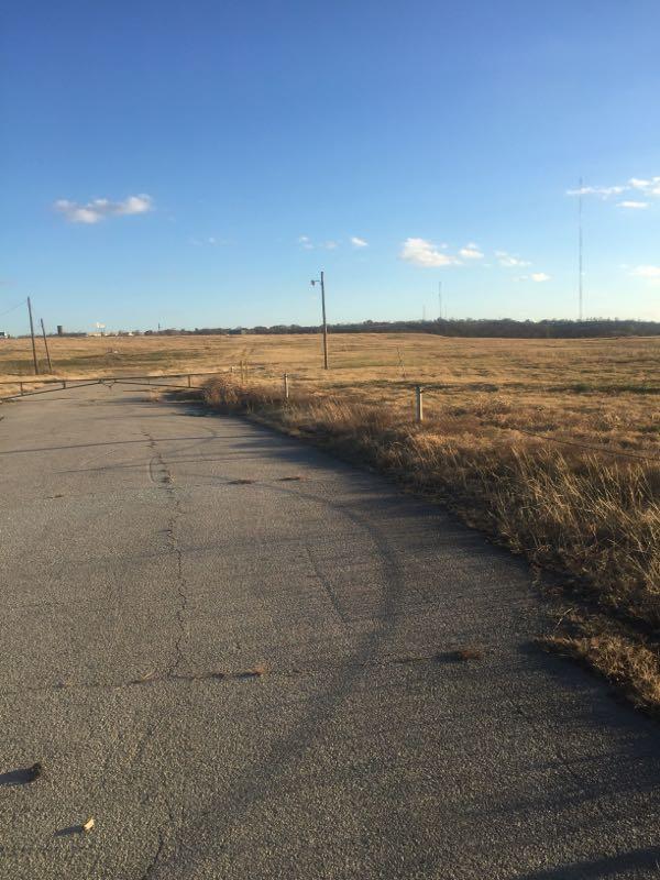 A curved, cracked asphalt road leads through a dry, open field under a clear blue sky. Sparse grass and utility poles line the edges of the road, which extends toward the horizon. Gateway Park mountain bike trail.