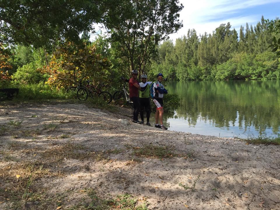 Three cyclists stand by a calm river surrounded by trees and greenery. They are wearing helmets and cycling gear, smiling at the camera. In the background, several bicycles can be seen resting on the ground. The setting features sandy terrain and reflective water, creating a serene outdoor atmosphere. Oleta River State Park mountain bike trail.