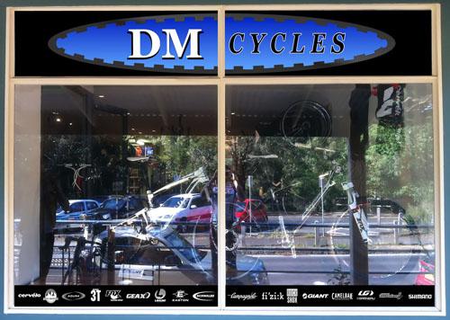 A storefront window displaying the name "DM CYCLES" in bold letters. The window showcases various bicycles and cycling gear inside the shop. A reflection of the exterior environment is visible in the glass.