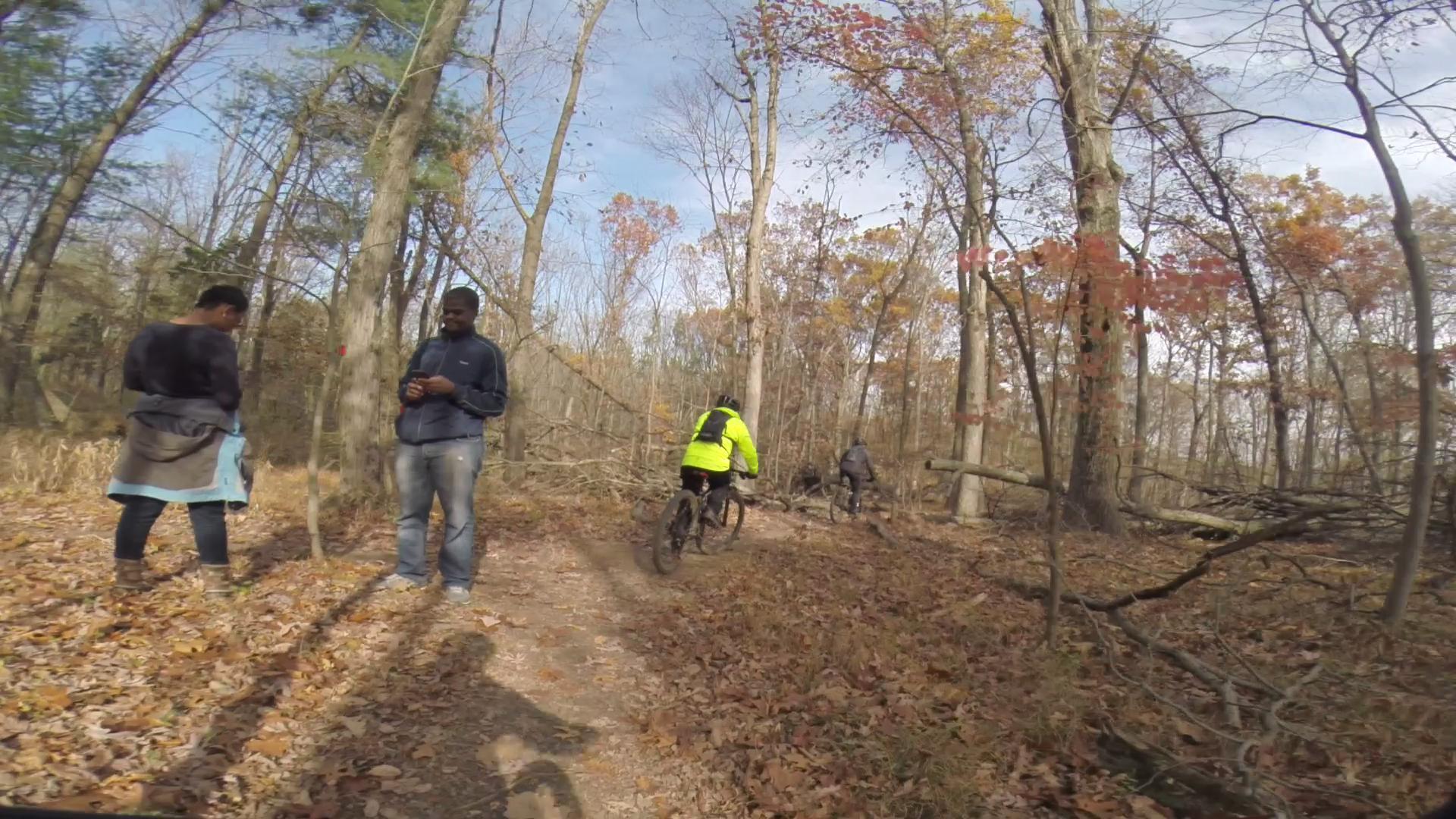 A group of people is enjoying a day in the woods. One person stands with their back turned, while another is looking at their phone. In the background, two mountain bikers ride along a dirt trail covered with fallen leaves, surrounded by trees with autumn foliage. The sky is partly cloudy. Chimney Rock mountain bike trail.