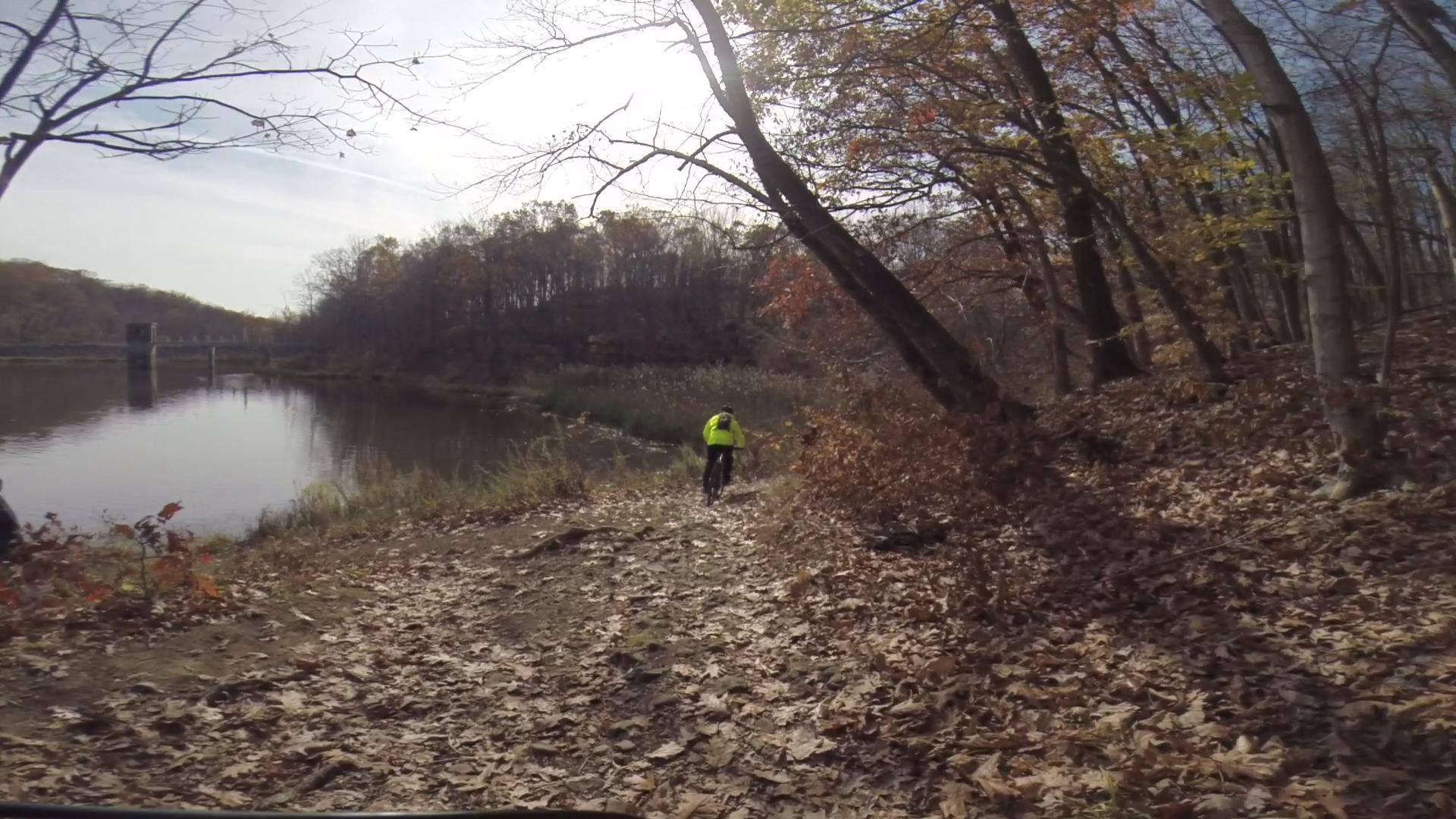 A person wearing a bright yellow jacket walks along a leaf-covered trail beside a calm body of water, surrounded by autumn foliage with trees in the background. The scene captures a peaceful outdoor setting in late fall. Chimney Rock mountain bike trail.