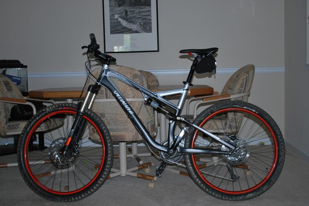 Specialized Stumpjumper FSR: A mountain bike with a silver frame and black handlebars, featuring orange accents on the wheels. The bike is positioned in an indoor setting with wooden furniture in the background, including a dining table and chairs.