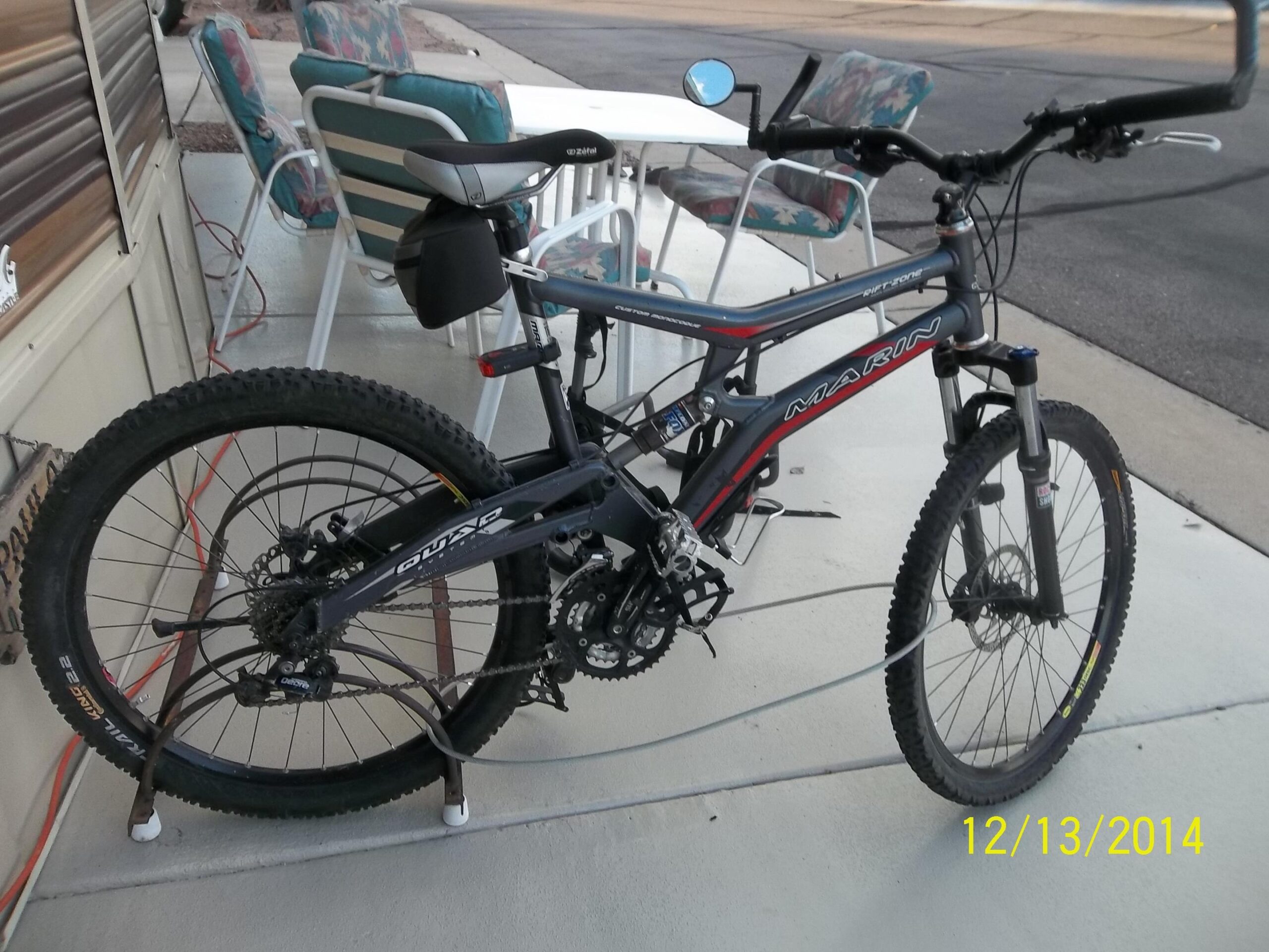 Marin Rift Zone: A gray Marin mountain bike is parked on a concrete surface next to a wall, secured in a bike rack. The bike features thick tires for off-road riding and is equipped with a rear bag. In the background, there are outdoor chairs with colorful fabric patterns and a visible road. A date is stamped in the lower right corner, reading 12/13/2014.