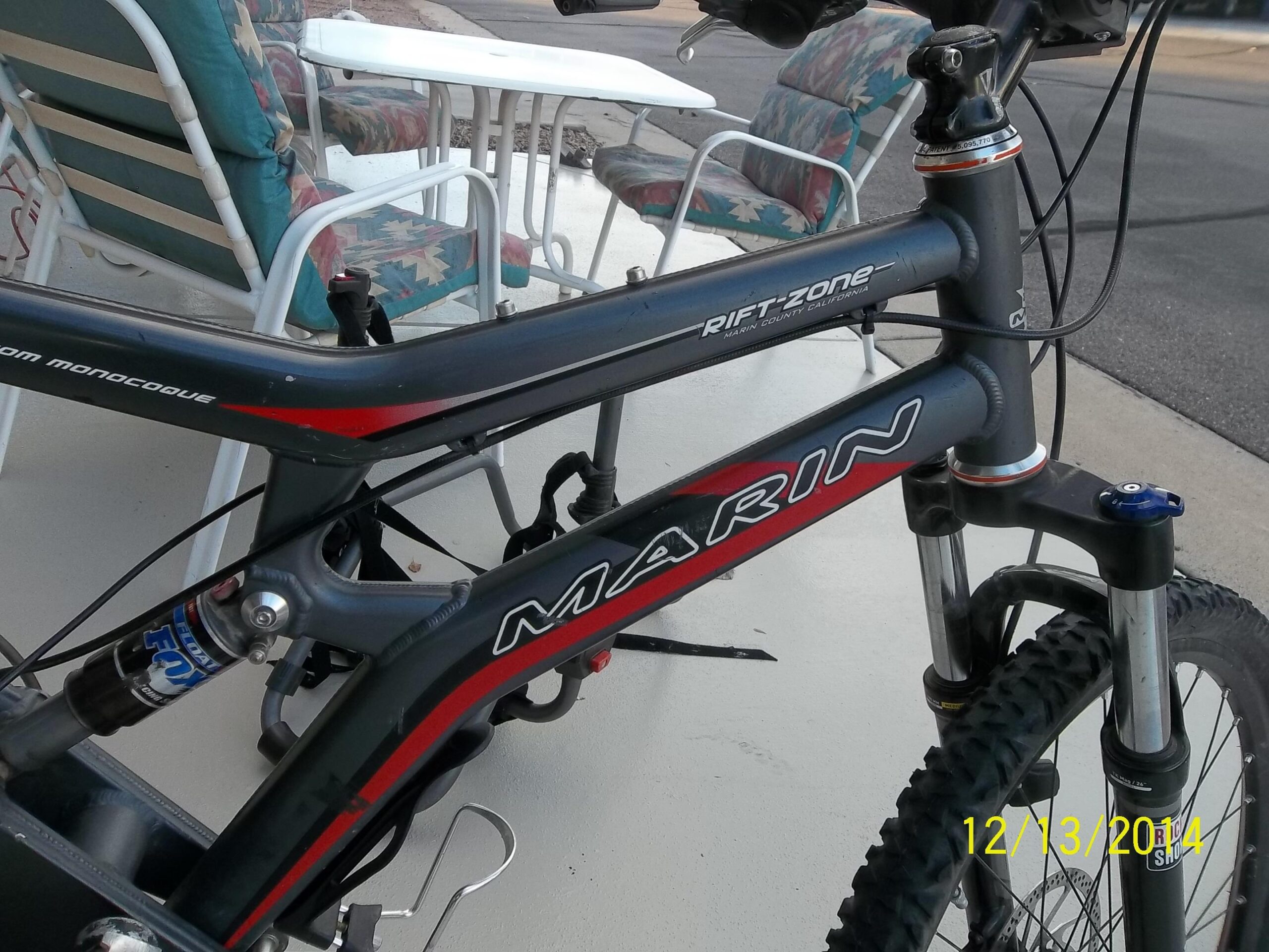 Marin Rift Zone: Mountain bike with a grey and red frame, featuring the Marin brand logo and the name "Rift Zone." The bike is positioned outdoors, near white patio furniture with colorful cushions. The date 12/13/2014 is displayed in the corner of the image.