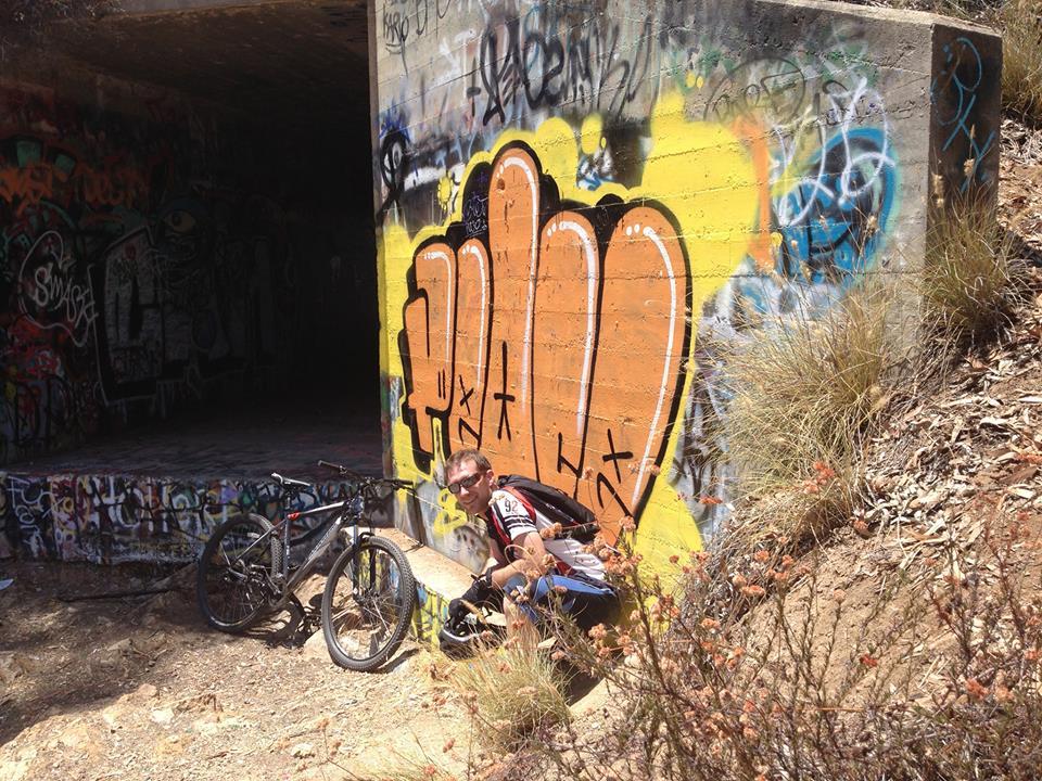 Marin Alpine Trail 29er: A person sitting on the ground next to a mountain bike, leaning against a brightly colored graffiti wall inside a tunnel. The area is surrounded by dry grass and small shrubs, with multiple layers of graffiti visible on the walls.