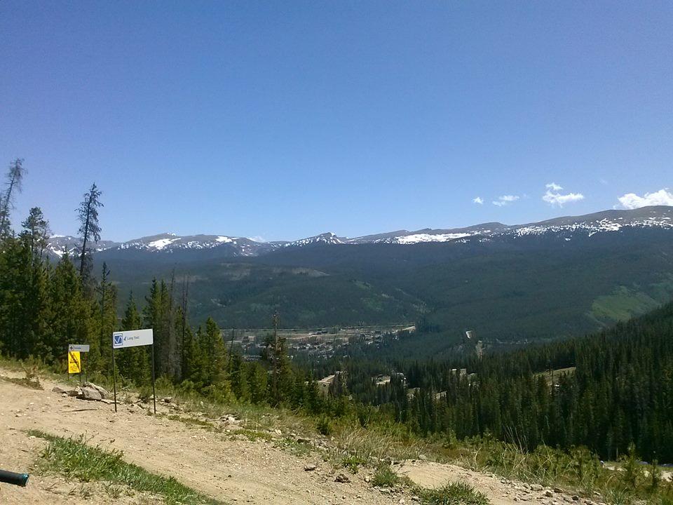 A panoramic view of a mountainous landscape featuring snow-capped peaks under a clear blue sky. The foreground shows a dirt path with signs indicating a trail or path, surrounded by lush green trees. In the distance, a small town is visible nestled in the valley. Trestle Bike Park mountain bike trail.