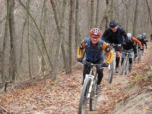 A group of mountain bikers navigating a dirt trail in a wooded area during autumn. The lead rider, wearing a red and orange jersey and a red helmet, is focused on the trail ahead, while others follow closely behind. The ground is covered with fallen leaves, and the trees around them are bare, indicating the season. Mountain Laurel Trails mountain bike trail.