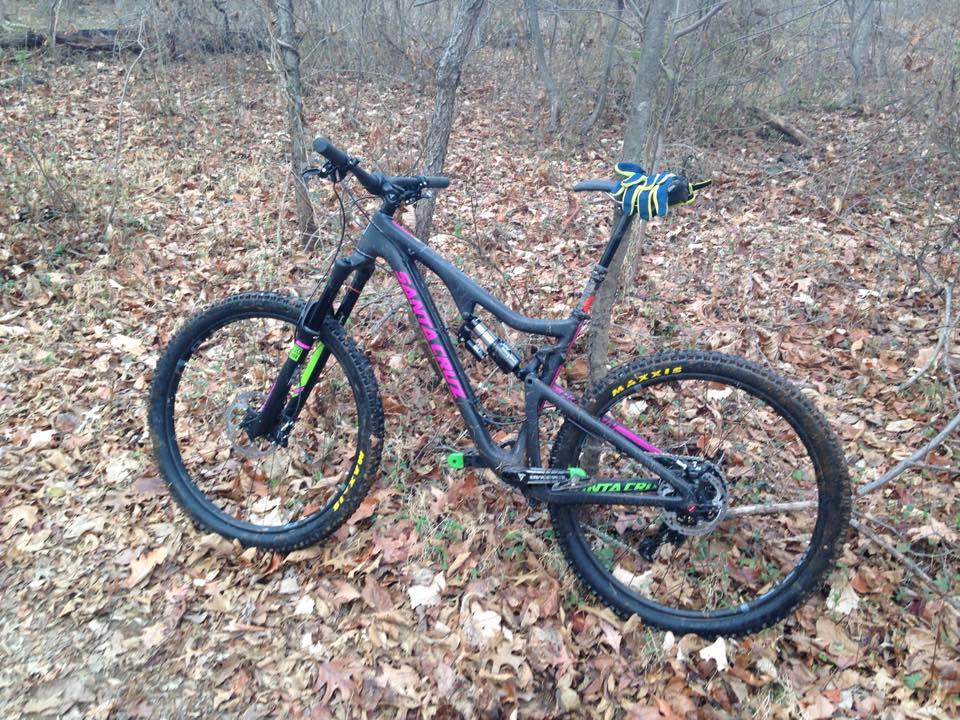 Santa Cruz Bronson: A mountain bike resting on a forest floor covered in autumn leaves. The bike features a sleek black frame with colorful accents in pink and green. It has wide tires suitable for off-road trails and a pair of gloves resting on the handlebars. Surrounding the bike are bare trees and scattered foliage, indicating a natural outdoor setting.
