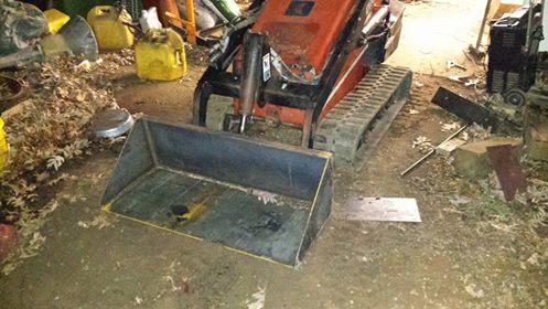 A small excavator with a bucket attachment parked in a cluttered shed, surrounded by tools and supplies, with leaves scattered on the ground. Mountain Laurel Trails mountain bike trail.