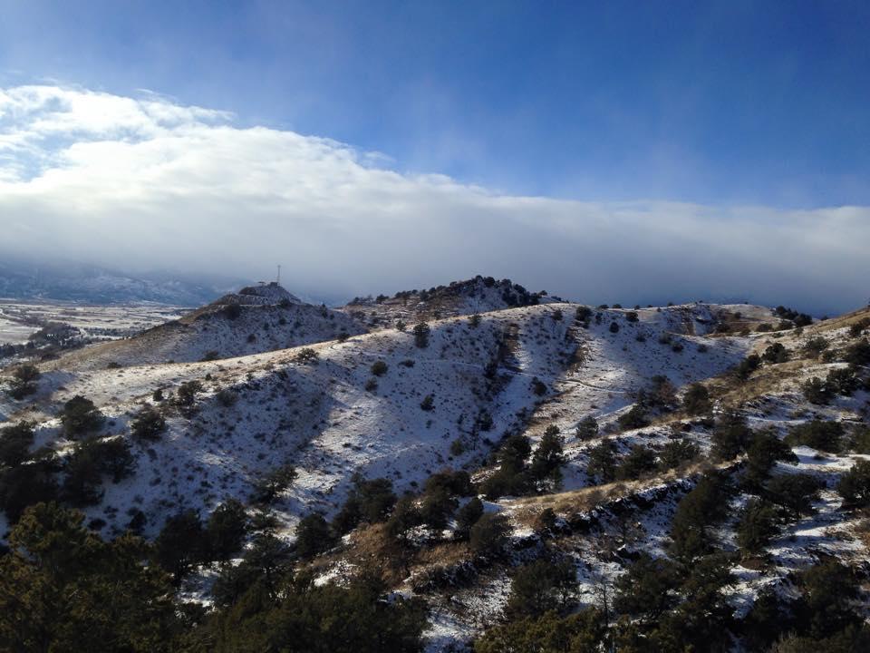 A scenic landscape featuring snow-covered hills under a blue sky with scattered clouds. The terrain is uneven, dotted with patches of evergreen trees, and a transmission tower is visible on one of the hills. Arkansas Hills mountain bike trail.
