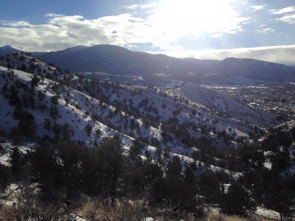 A scenic view of snow-covered hills and mountains under a bright sky, with patches of sunlight illuminating the landscape. The foreground features trees scattered throughout the slopes, and the distant mountains create a serene backdrop. Arkansas Hills mountain bike trail.