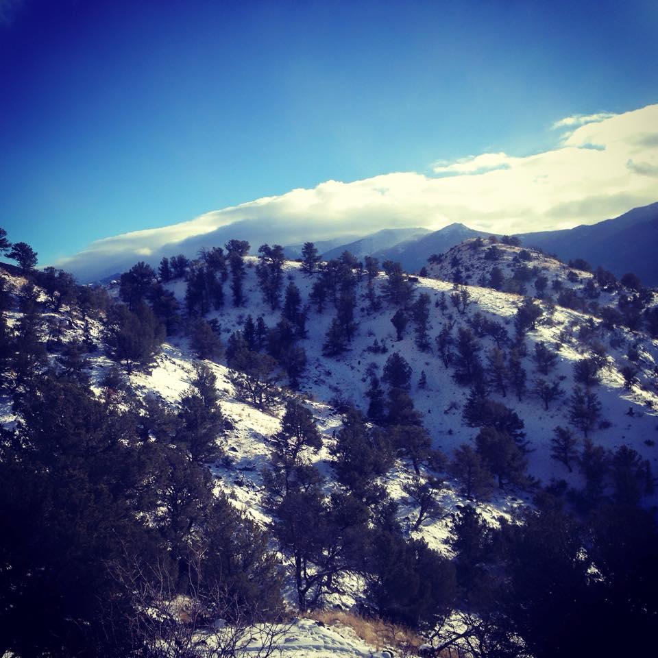 A scenic landscape featuring snow-covered hills and pine trees under a bright blue sky, with distant mountains partially obscured by clouds. Arkansas Hills mountain bike trail.
