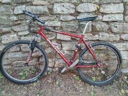 Specialized hardrock: A red mountain bike with black tires and a silver frame is leaning against a textured stone wall. The bike has a front suspension and is set on a dirt path with some greenery in the background.