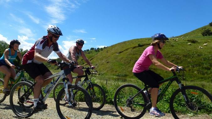 A group of four cyclists riding on a gravel path surrounded by lush green hills and a clear blue sky. The cyclists, wearing helmets and casual athletic clothing, appear to be enjoying a sunny day outdoors. Clutha Gold Trail mountain bike trail.